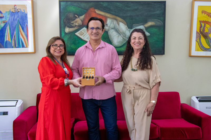 Tr&ecirc;s pessoas posam para uma foto em uma sala, diante de um sof&aacute; vermelho e obras de arte. Ao centro, um homem segura uma pequena pe&ccedil;a decorativa, ladeado por duas mulheres sorridentes.