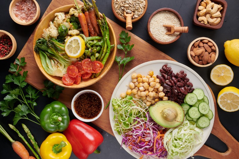 Pratos com vegetais, gr&atilde;o-de-bico, feij&atilde;o, abacate e pepino, acompanhados de castanhas, sementes e piment&otilde;es coloridos, dispostos sobre uma mesa de madeira