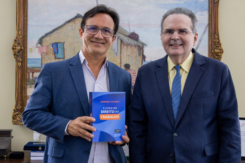 Dois homens de terno posam lado a lado; o da esquerda segura um livro azul intitulado “Curso de Direito do Trabalho”, diante de uma pintura emoldurada