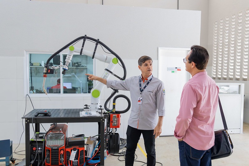 Homem aponta para um equipamento de soldagem robotizada enquanto conversa com outro homem em laborat&oacute;rio. Ao fundo, h&aacute; bancadas, m&aacute;quinas e outros equipamentos utilizados em atividades de pesquisa