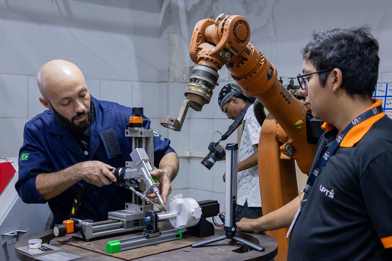 Homem opera equipamento em bancada enquanto outro acompanha o trabalho. Ao fundo, um braço robótico industrial e um terceiro homem com câmera completam a cena em laboratório de pesquisa